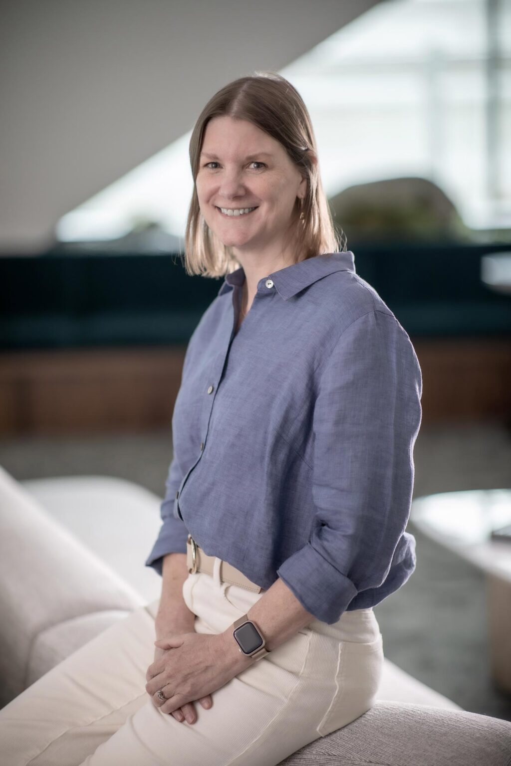 Sandra Beer, Senior Project Leader at GFF, seated on a light-colored sofa wearing a blue blouse and white pants, smiling in a modern office interior.