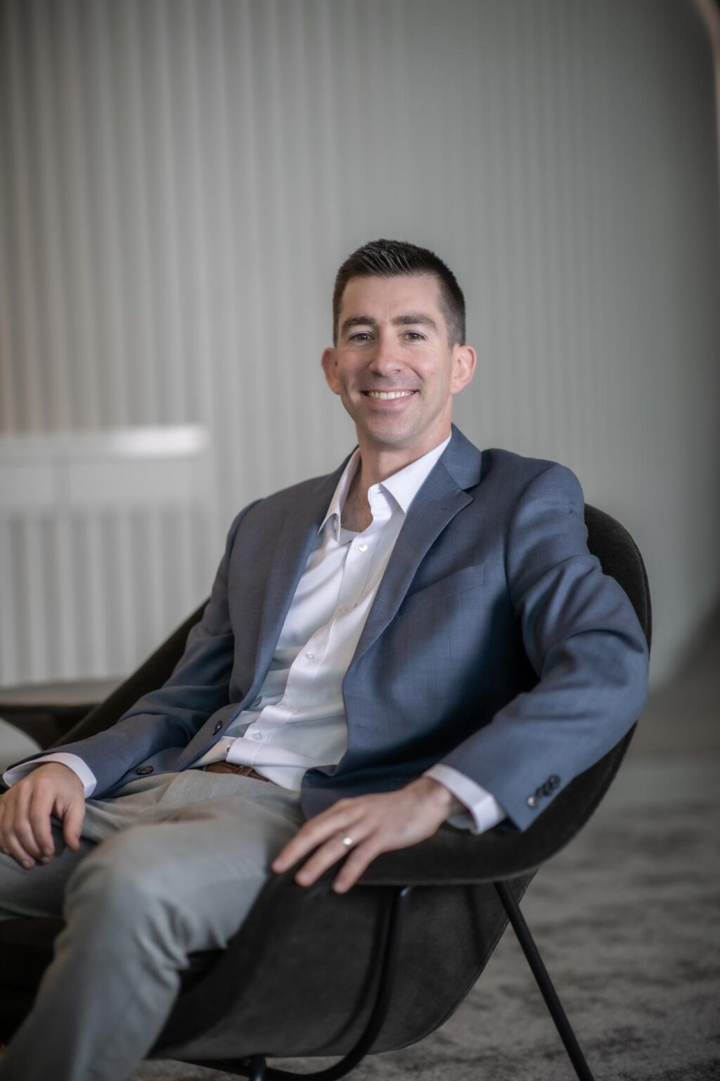 A portrait of Adam Fenner, Project Leader at GFF, seated in a modern chair wearing a light blue blazer and white shirt, smiling at the camera.