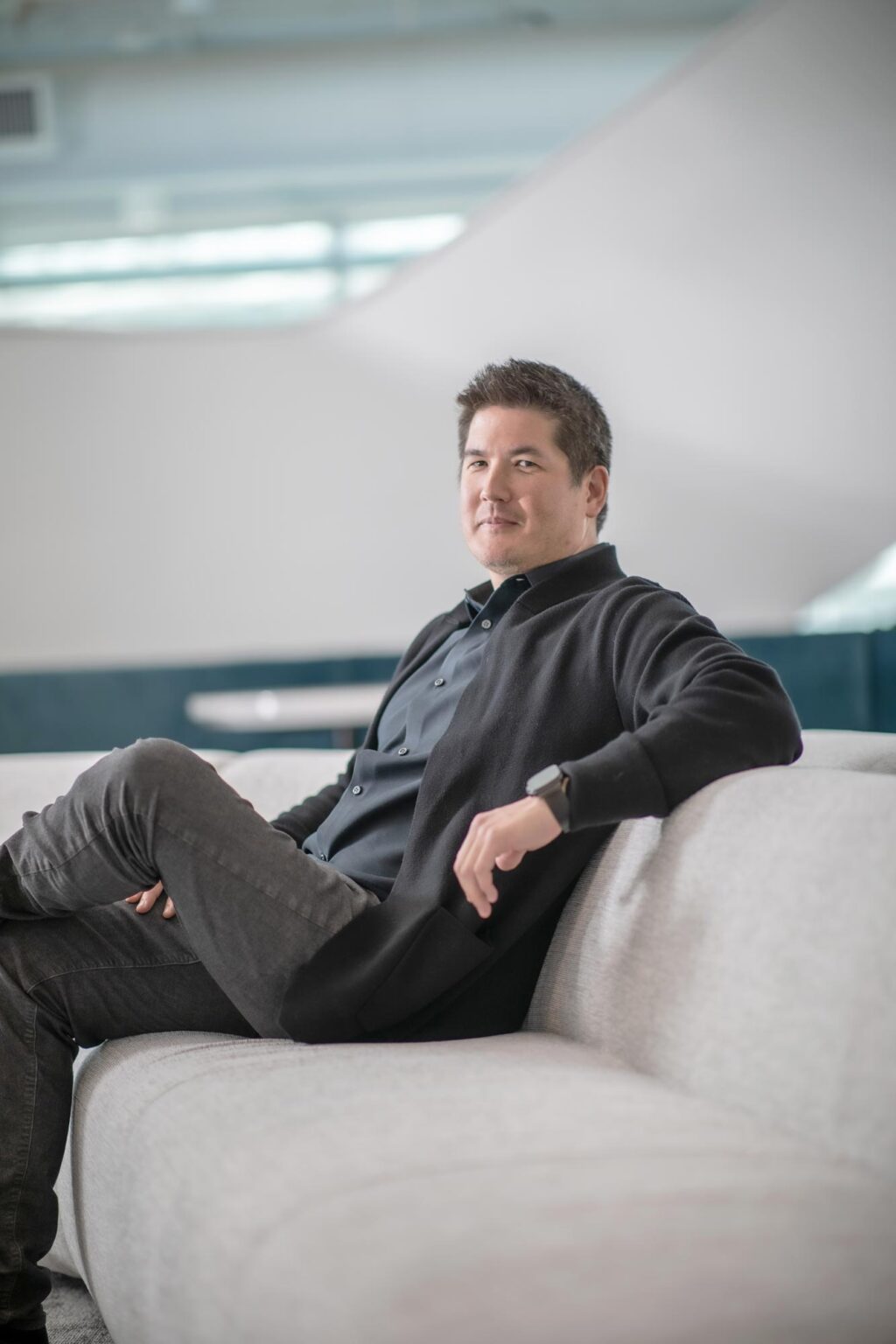 David Colwill seated on a light gray sofa in a modern office interior, wearing a dark jacket and relaxed expression.