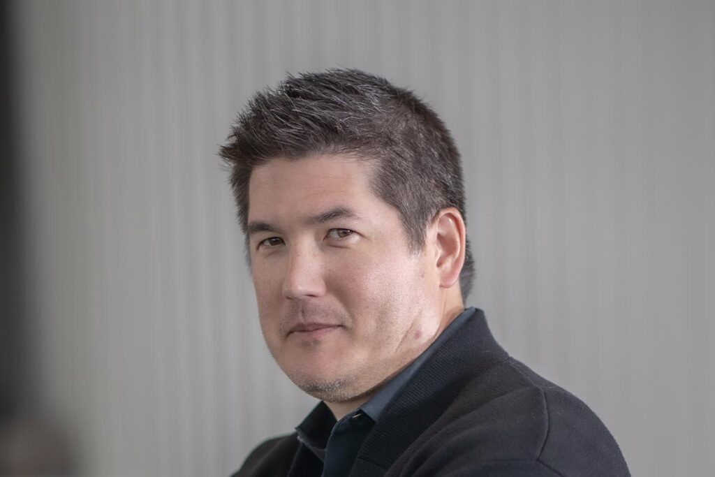 Close-up portrait of David Colwill wearing a dark shirt and jacket, with a neutral blurred background and soft natural light.
