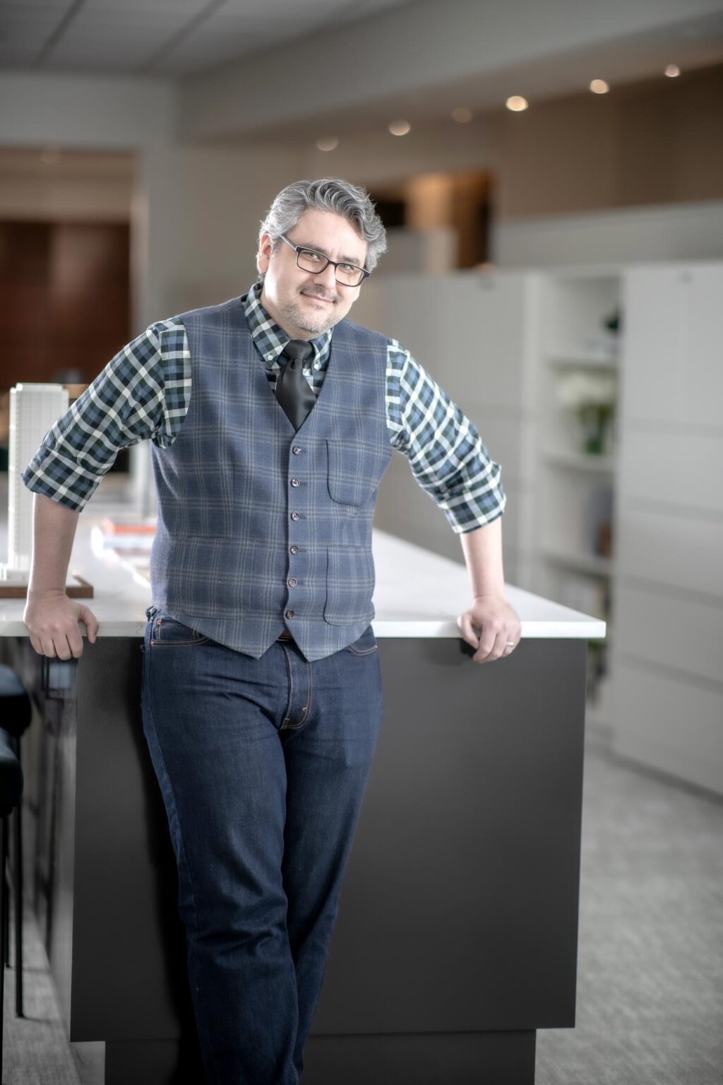 Jason Fedors leaning against a countertop in a modern office, wearing a plaid shirt, tie, and vest, smiling confidently.