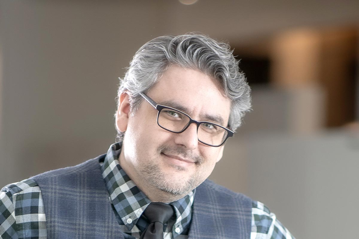 Close-up portrait of Jason Fedors, wearing glasses and a plaid shirt with a vest, smiling slightly in a softly lit office interior.