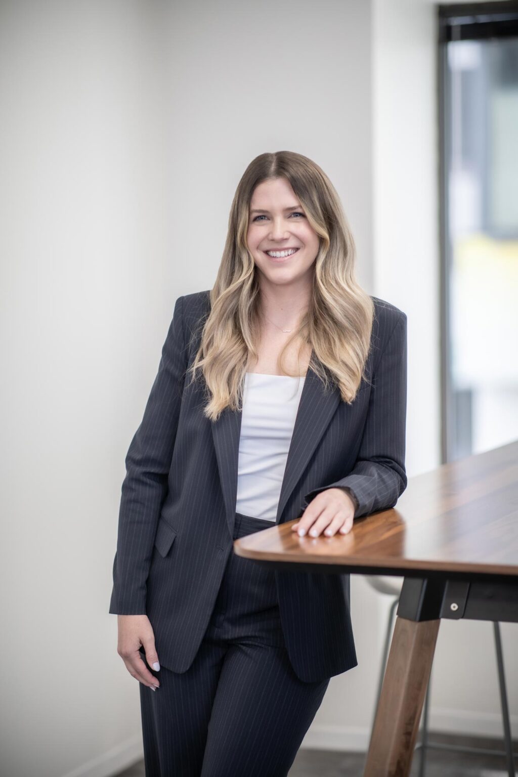 A portrait of Jenna Ross, Interior Design Leader at GFF, standing beside a tall wood table in a modern office setting, wearing a dark pinstripe suit.