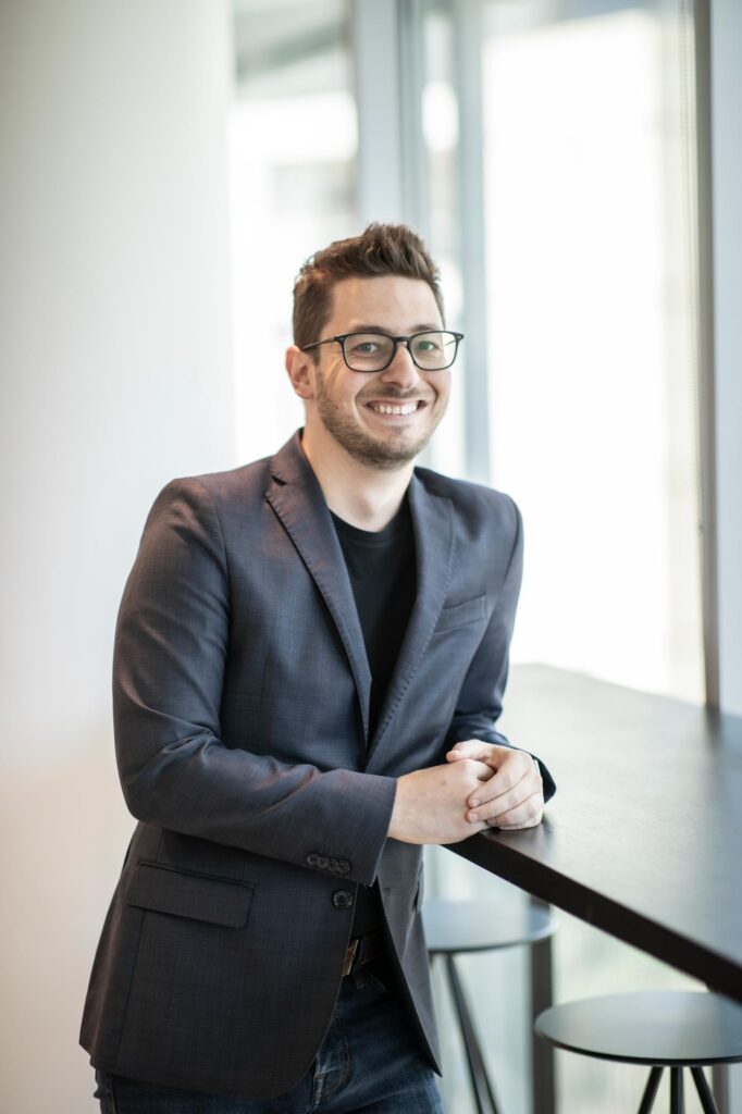 Josh Hafel leaning casually at a tall counter in a bright office space, smiling and wearing glasses with a dark blazer.