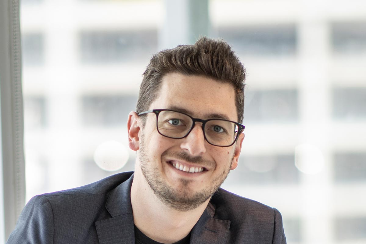 Close-up portrait of Josh Hafel smiling and wearing glasses and a dark blazer in a bright office setting.
