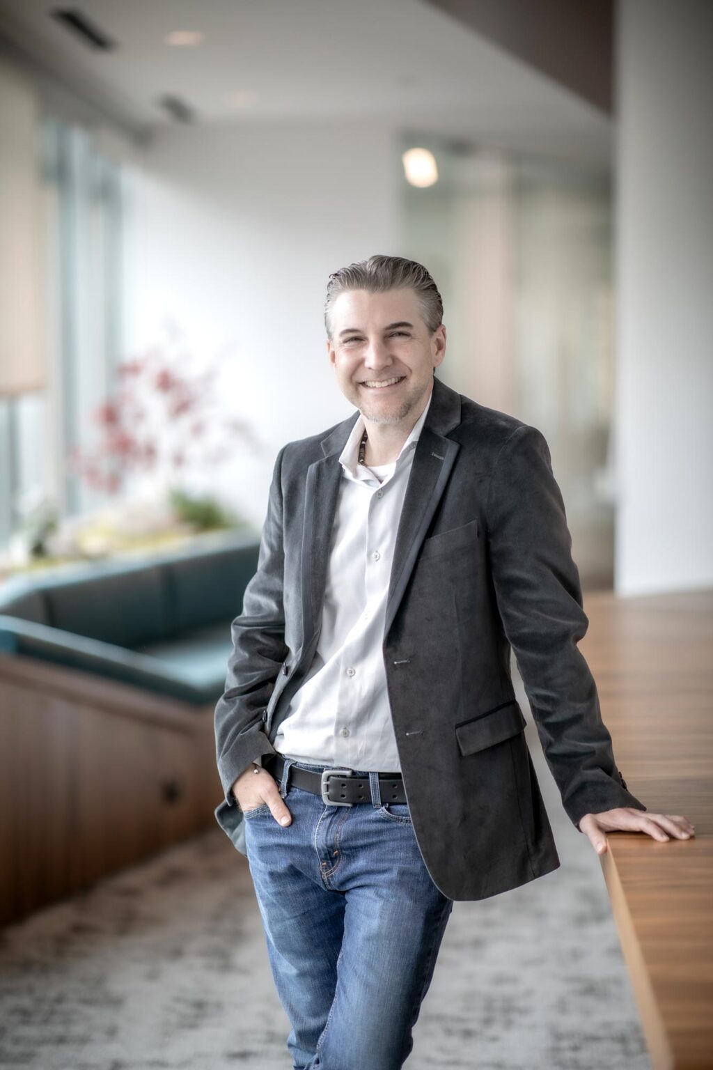 A portrait of Rocky Owens, Project Leader at GFF, leaning casually against a wood countertop in a bright modern office interior, wearing a dark jacket and jeans.