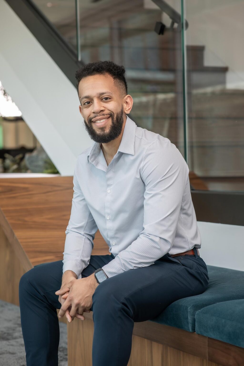 A portrait of Thomas Castro, Project Leader at GFF, seated on a wooden bench in a modern office interior with glass and metal details, smiling while wearing a light gray shirt and dark trousers.