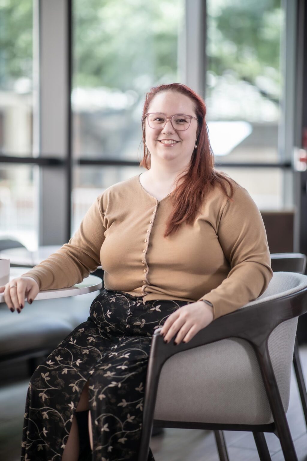 Alexis Kaleri seated in a bright interior with large windows, wearing a tan sweater and patterned skirt, smiling toward the camera.