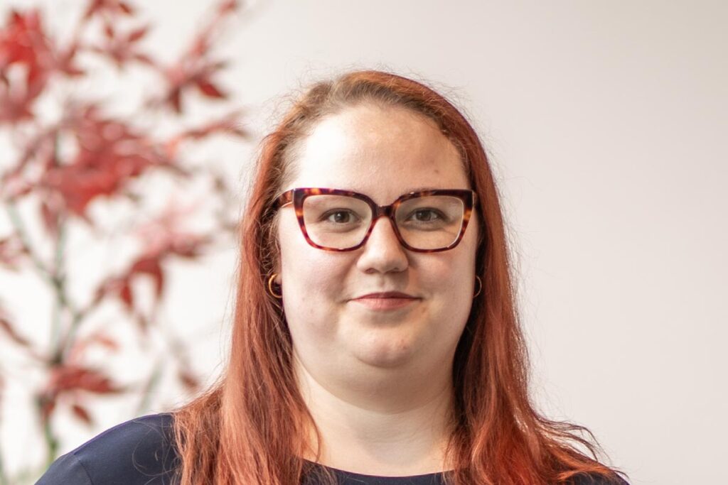 Close-up portrait of Alexis Kaleri wearing glasses and a navy top, smiling softly with a blurred background of red leaves.