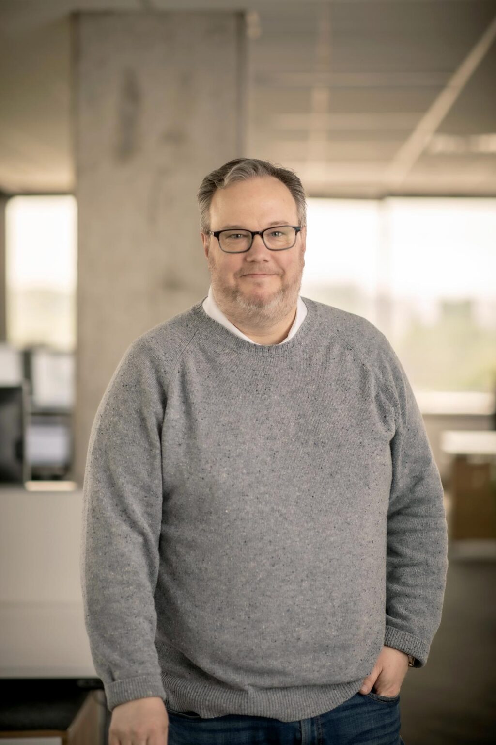 Chad Kyzar standing in a bright office interior with natural light and concrete elements, wearing a gray sweater and jeans.