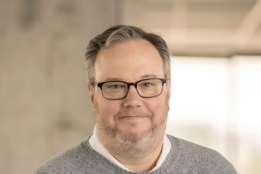 Close-up portrait of Chad Kyzar wearing glasses and a gray sweater, in front of a softly blurred office setting.