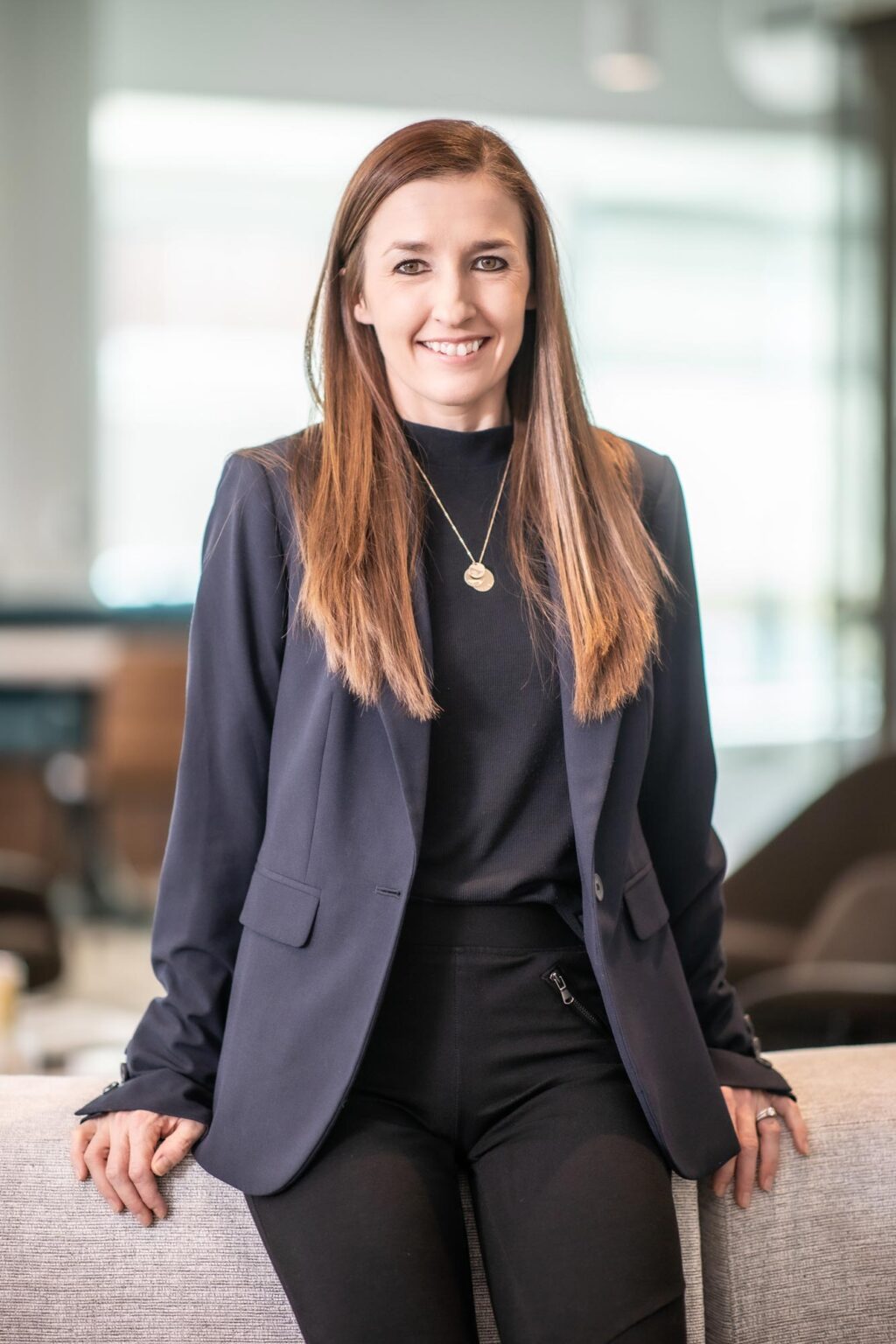 Heather Abaya seated on the back of a light gray sofa in a modern office, smiling and wearing a dark blazer and pendant necklace.