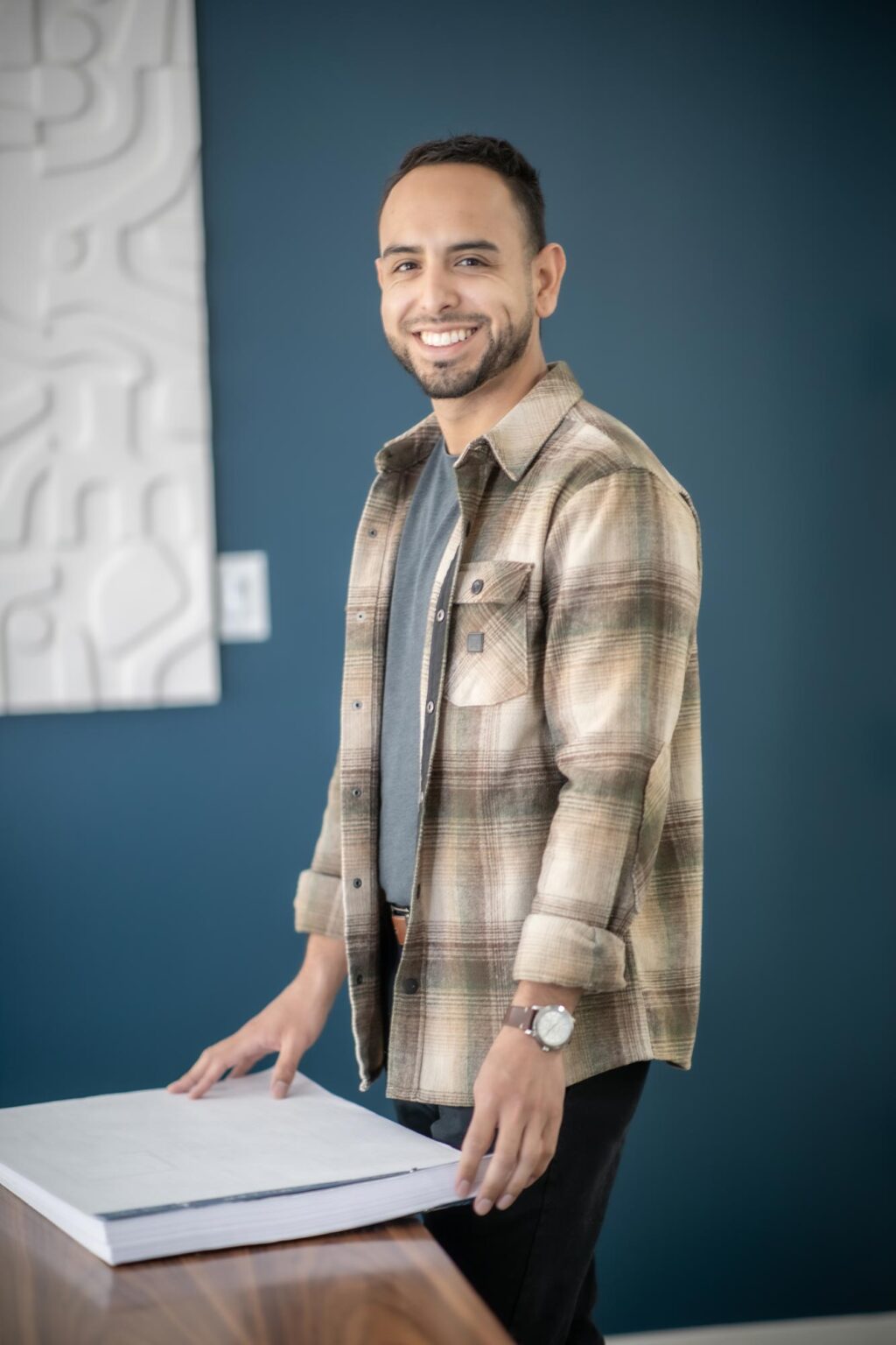Jake Chavez standing at a table with architectural drawings in a modern office, wearing a plaid shirt and smiling.