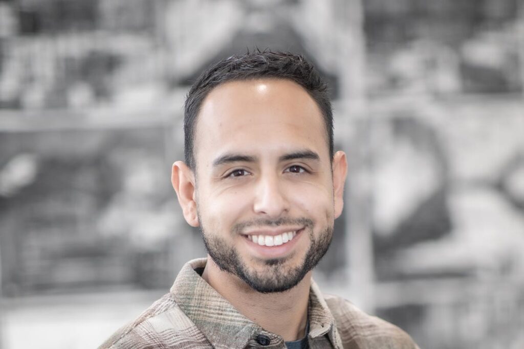 Close-up portrait of Jake Chavez wearing a plaid shirt, smiling in front of a softly blurred architectural background.