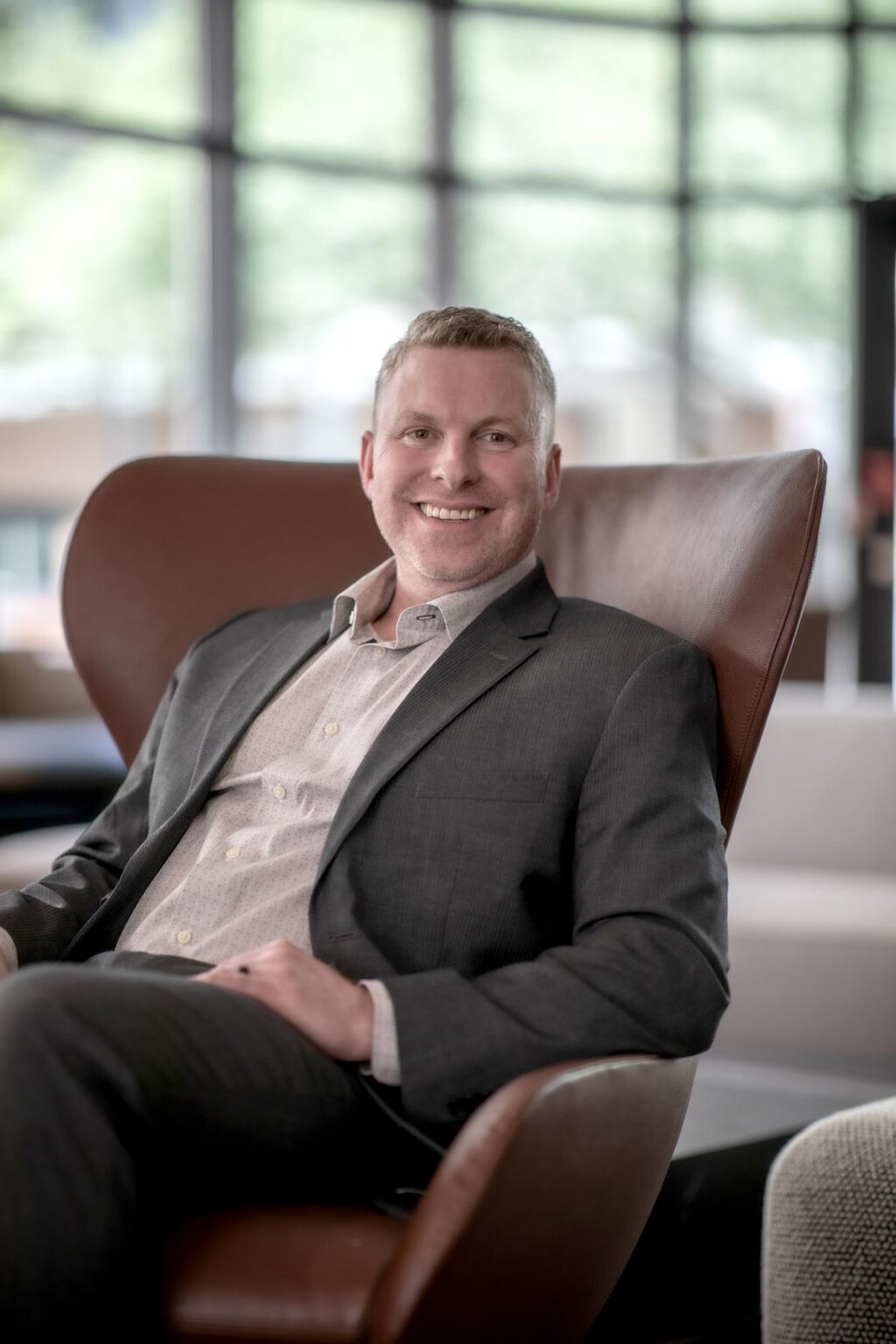 A portrait of Joshua Brown, Senior Project Coordinator at GFF, seated in a modern office interior with large windows, wearing a dark gray suit and light patterned shirt.