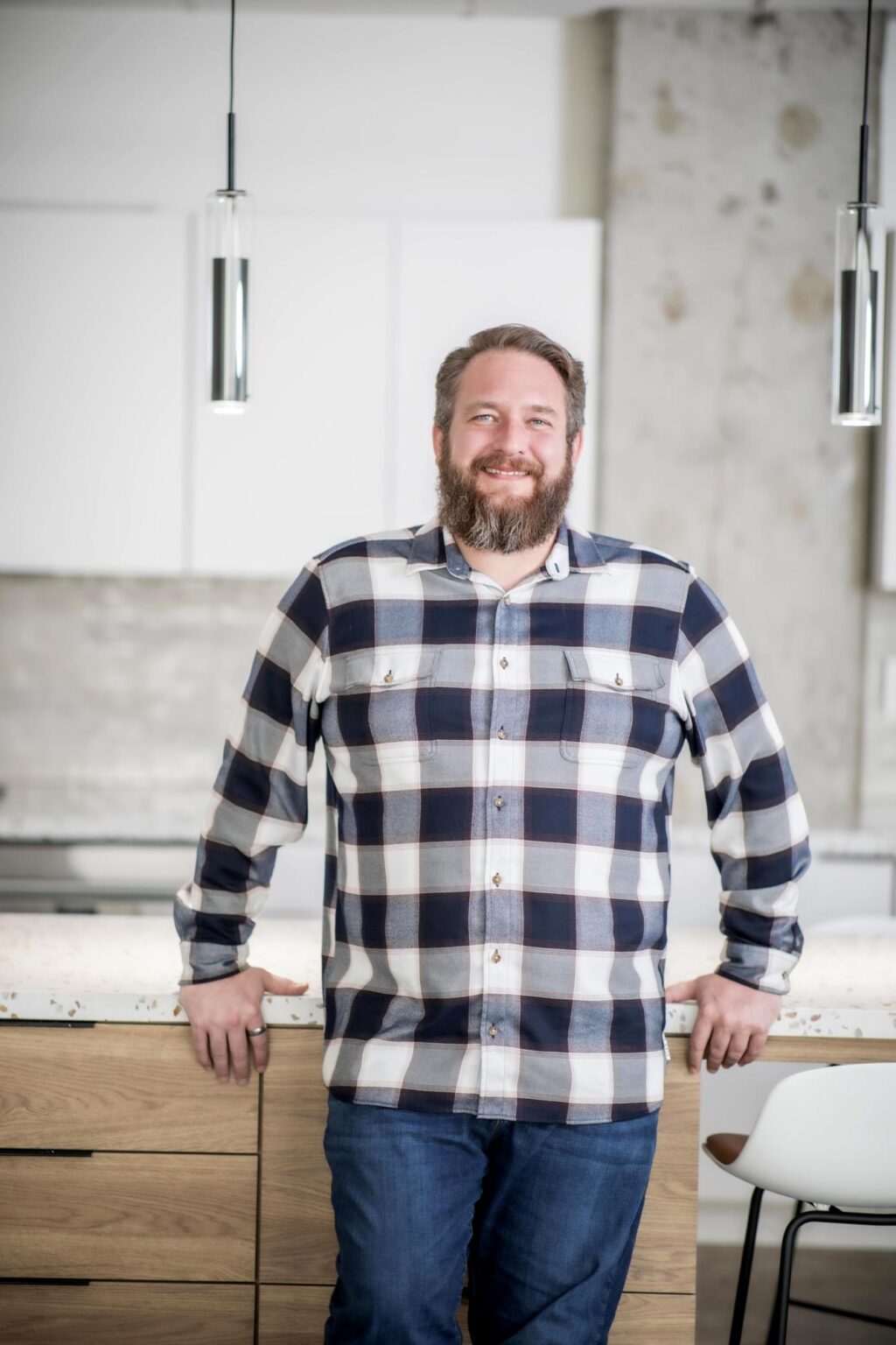 A portrait of Scott Williams, Senior Project Coordinator at GFF, standing in a bright modern office kitchen wearing a plaid shirt and jeans, smiling with hands resting on the countertop.