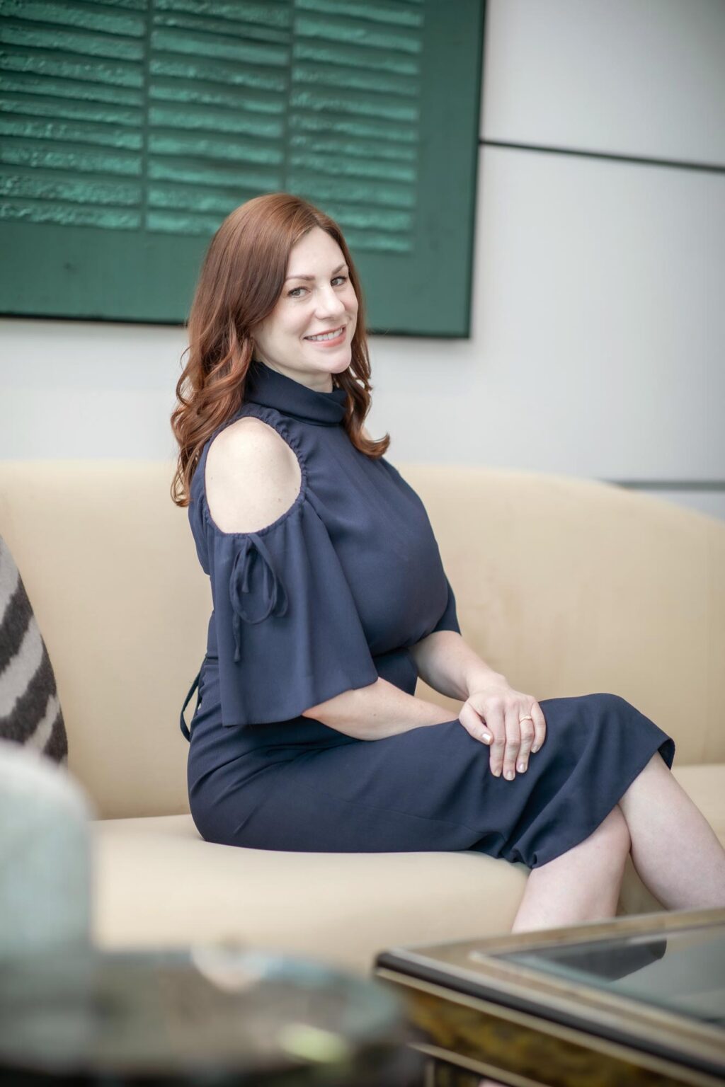 Shannon Fahey seated on a beige sofa wearing a navy dress, smiling and looking toward the camera in a modern interior space.