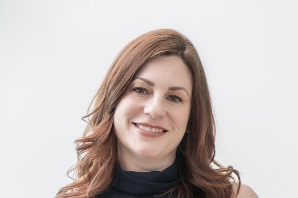 Close-up portrait of Shannon Fahey smiling with softly curled hair, wearing a navy dress against a white background.