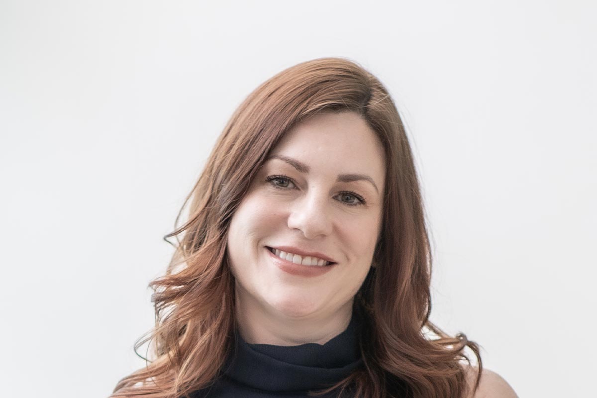 Close-up portrait of Shannon Fahey smiling with softly curled hair, wearing a navy dress against a white background.