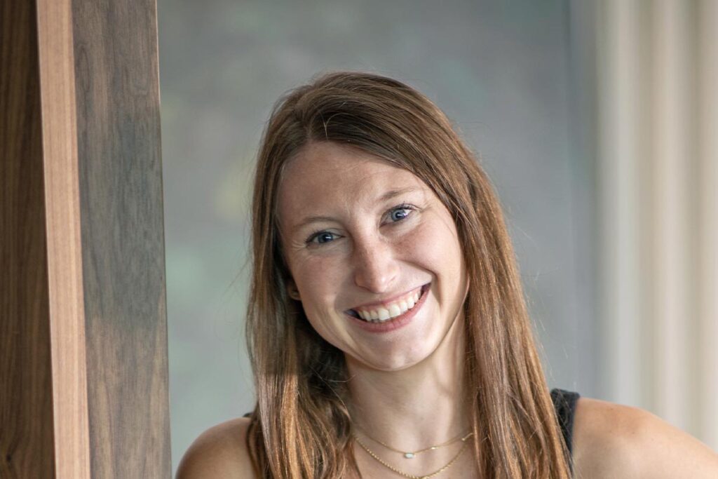 Close-up portrait of Taylor Wade standing beside a wood panel, wearing a black dress and gold necklaces, with a softly blurred interior background.