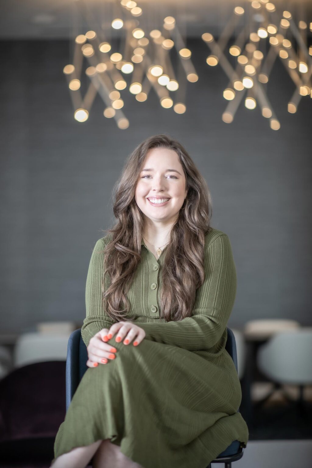 Camille Vigil, NCARB RA, Project Coordinator at GFF, seated under modern pendant lighting in a green dress, photographed in a contemporary office environment.
