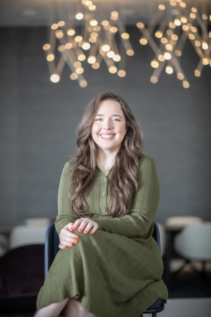 Camille Vigil, NCARB RA, Project Coordinator at GFF, seated under modern pendant lighting in a green dress, photographed in a contemporary office environment.