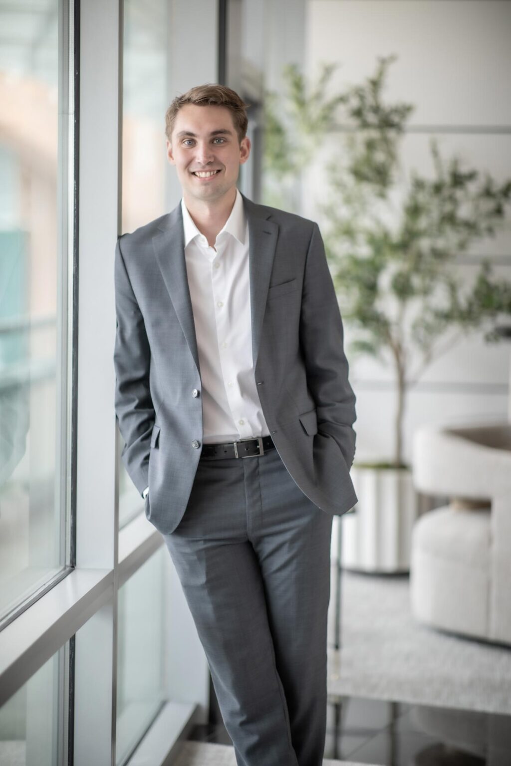 Carson Guy, Project Coordinator at GFF, standing by a window in a gray suit and white shirt with greenery in the background.
