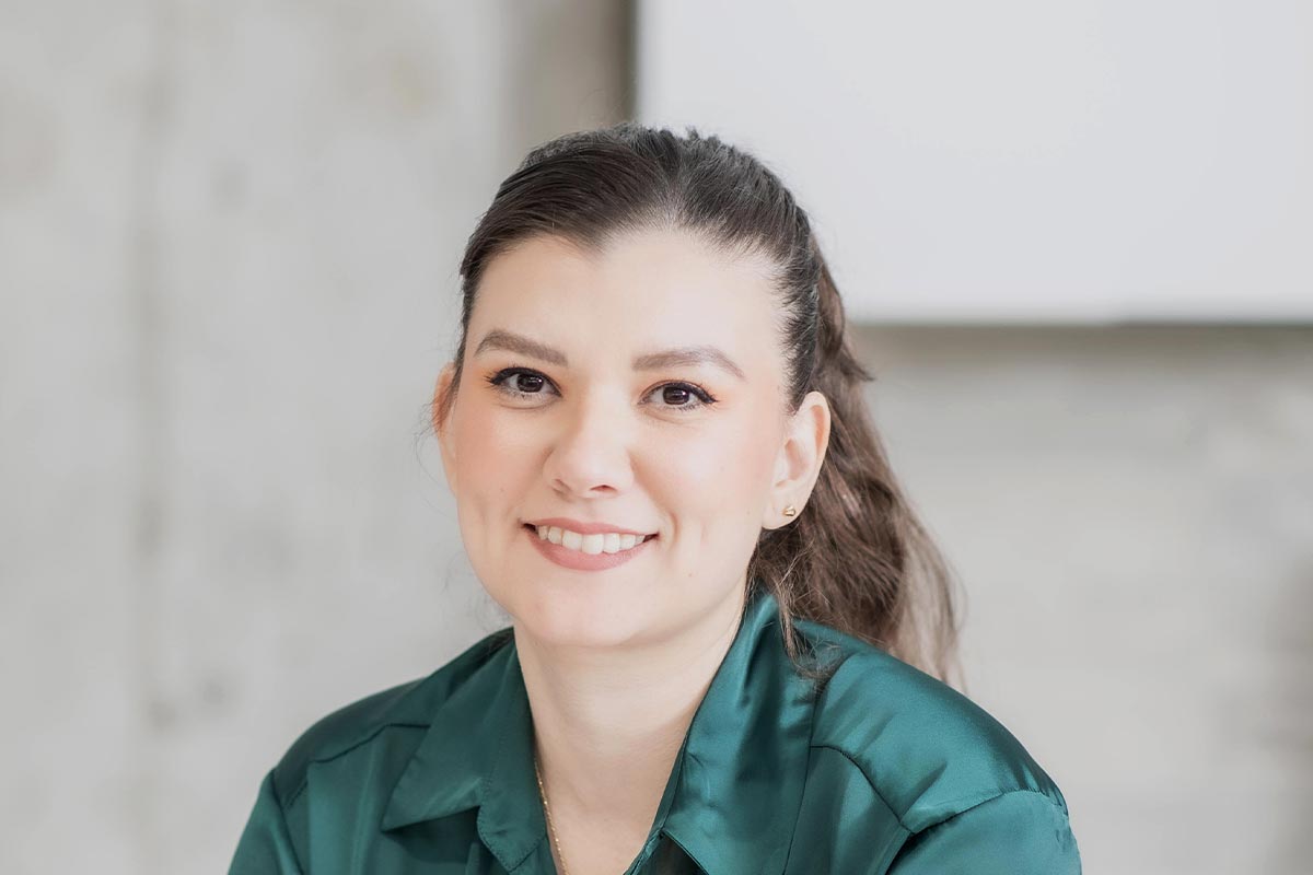 Close-up portrait of Cecilia Garcia wearing a green blouse, smiling in a light-filled contemporary office setting.
