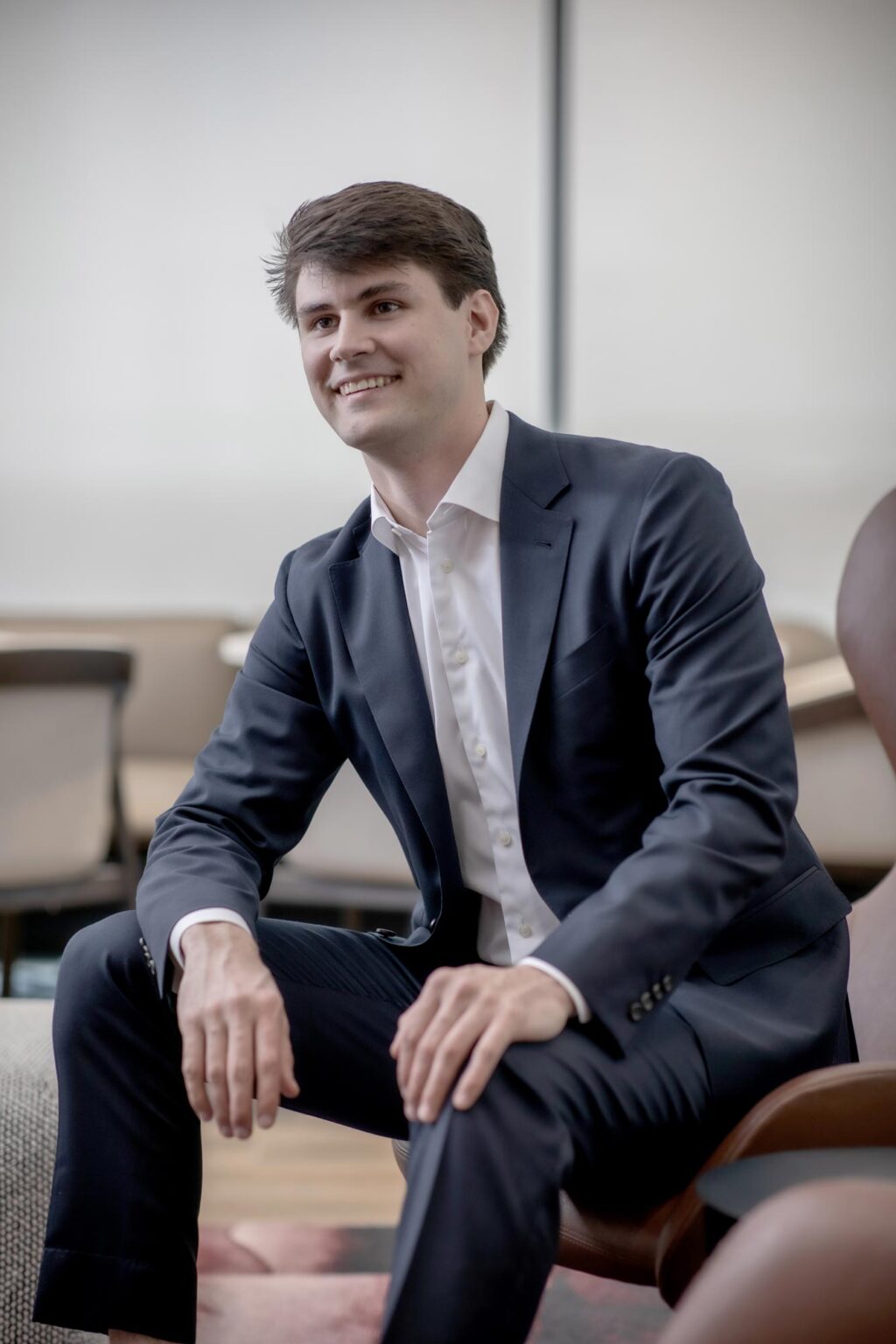 Coleman Brink, Design Coordinator at GFF, seated in a modern office chair wearing a navy suit and white shirt, smiling in a light-filled workspace.