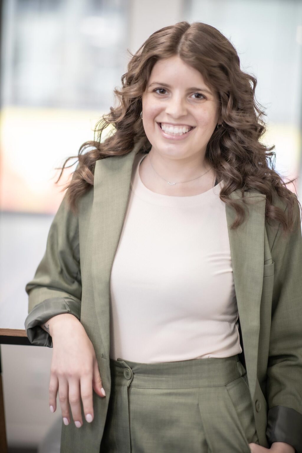 A portrait of Gabrielle Duran, Project Coordinator at GFF, standing in the Austin office wearing an olive green jacket and light top, smiling with one hand resting on a railing.