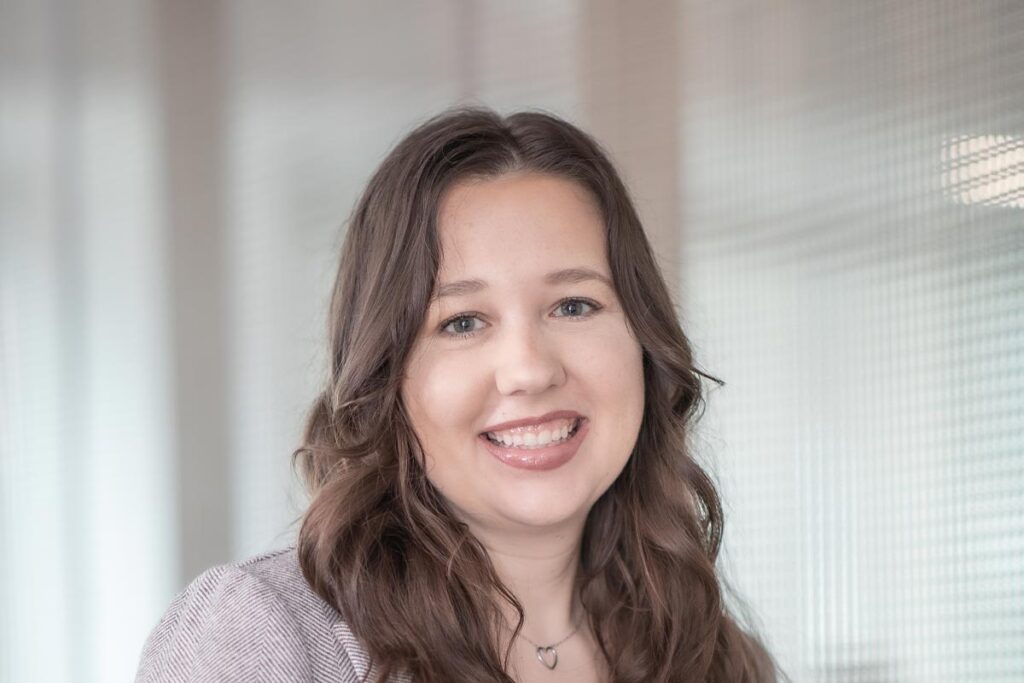 Close-up portrait of Kelsey Kohlmeier wearing a light gray blazer and white blouse, smiling in a softly lit office setting.