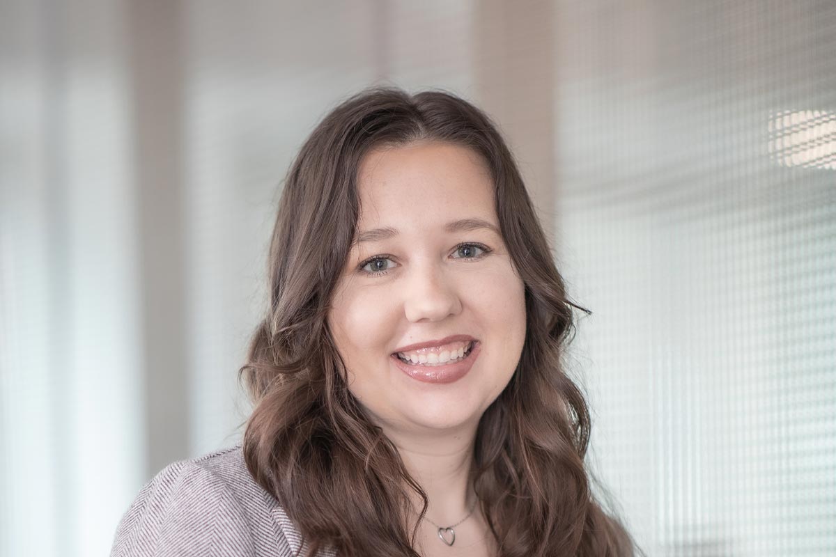 Close-up portrait of Kelsey Kohlmeier wearing a light gray blazer and white blouse, smiling in a softly lit office setting.