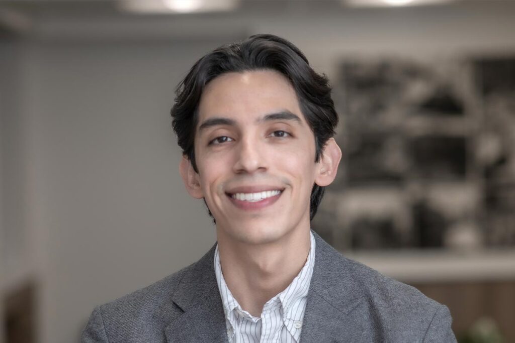 Close-up portrait of Woody Owens wearing a gray jacket and striped shirt, in front of a neutral office backdrop.