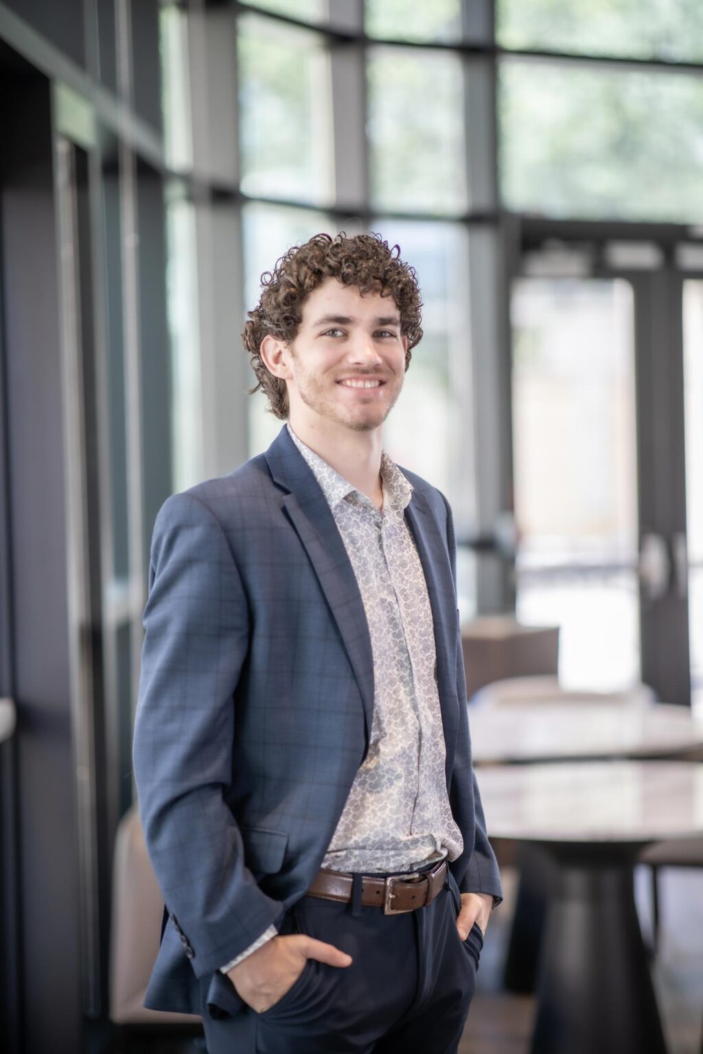 A portrait of Jonathan Moebius, Design Professional at GFF, standing in a bright glass-lined interior with hands in pockets, wearing a patterned shirt and blue blazer.