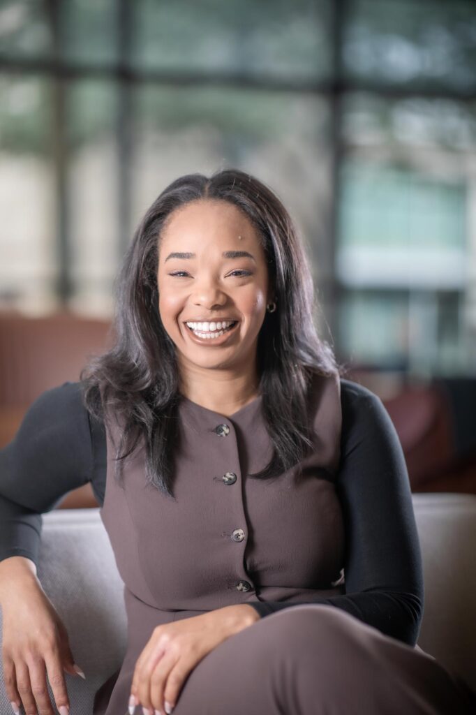 A portrait of Leah Coley, Architectural Professional at GFF, seated in a modern office interior with large windows, smiling and wearing a brown dress with black sleeves.