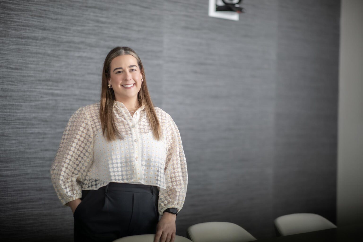 Maya Pavon, Design Professional at GFF, standing in a modern conference room wearing a sheer patterned blouse and dark trousers.