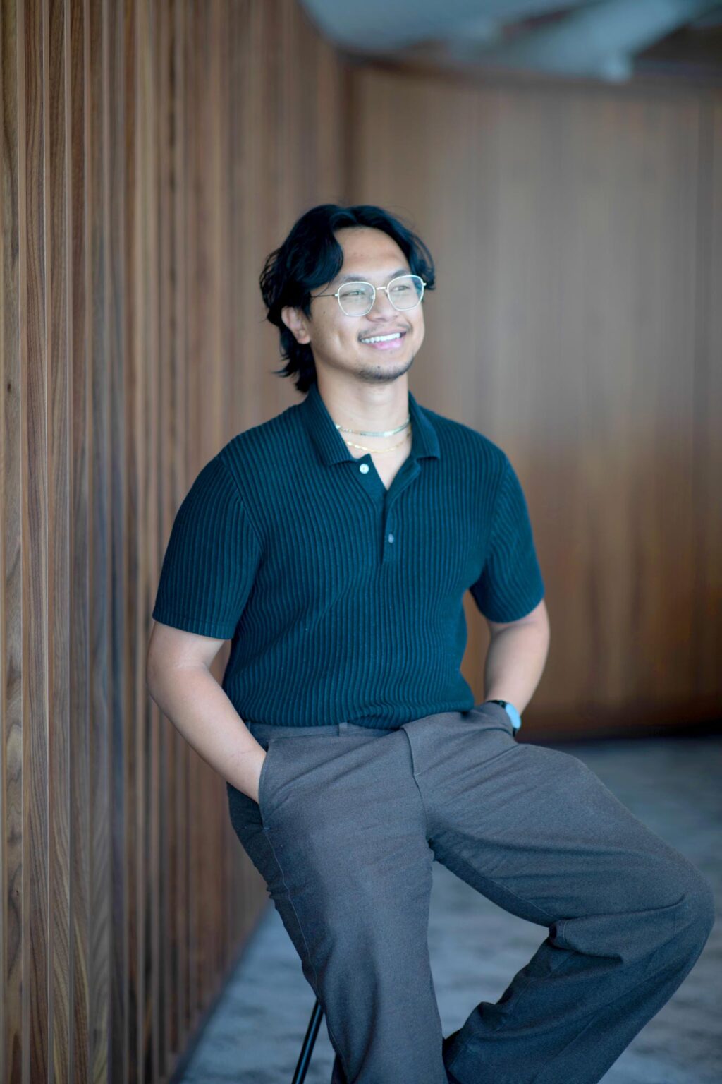Vince Hernandez, Architectural Professional at GFF, seated against a wood-paneled wall in the Austin office, wearing glasses and a dark shirt, photographed in a contemporary design studio environment.