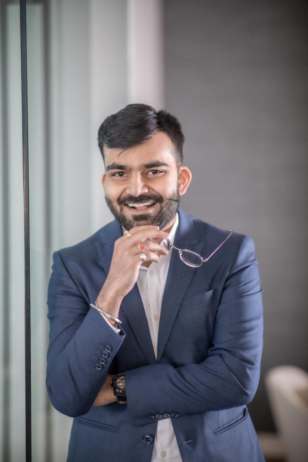 Nikunj Vara, Architectural Professional at GFF, smiling in a navy suit while holding eyeglasses, photographed in a modern design studio environment.