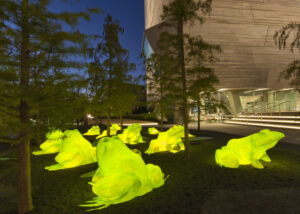 An evening photograph of Illuminated frog sculptures sitting in a landscape area adjacent to the entrance to the Perot Museum of Nature & Science