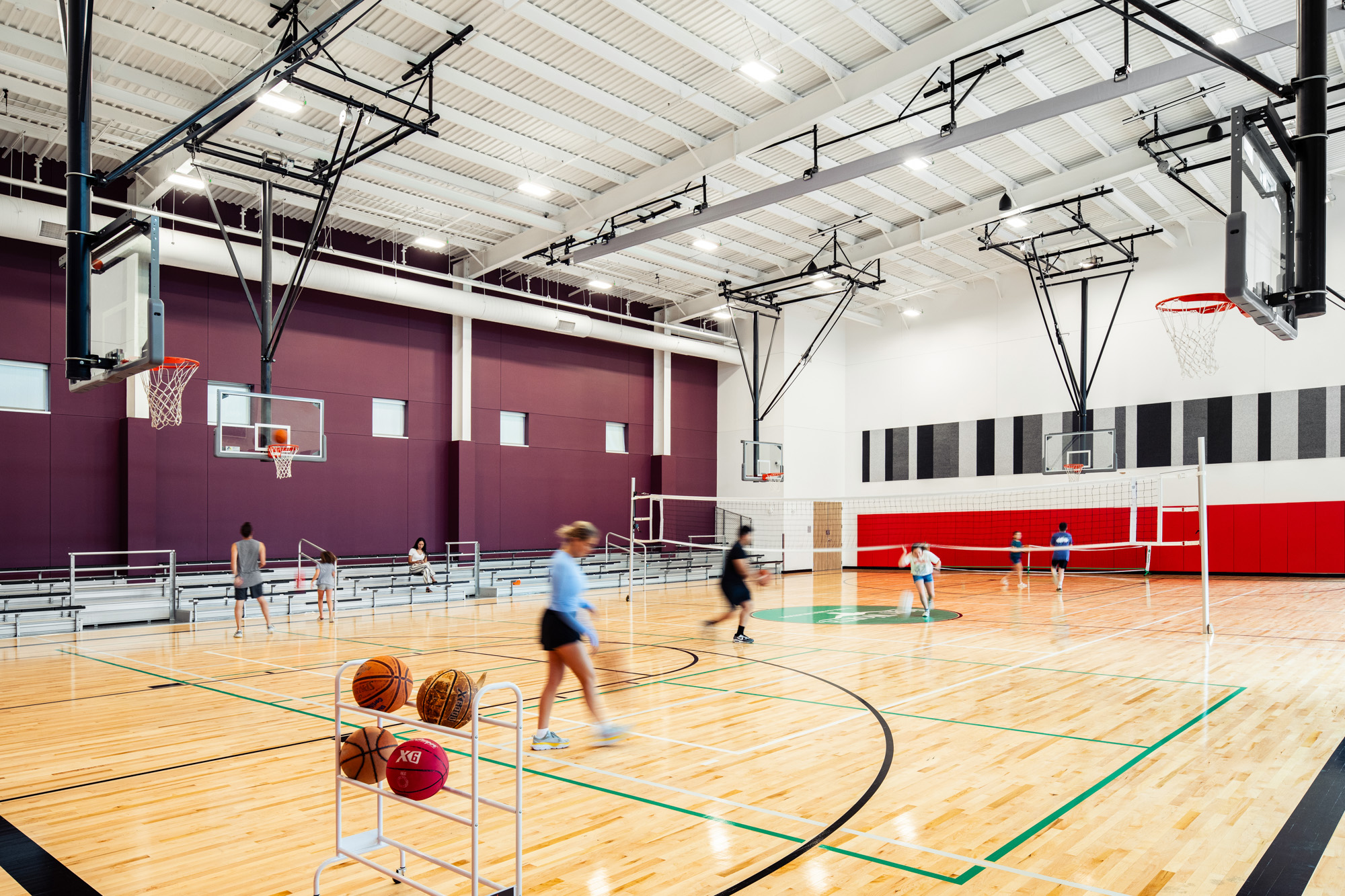 A wide view of a competition-sized gymnasium with basketball and volleyball courts, featuring bleachers and players in action.