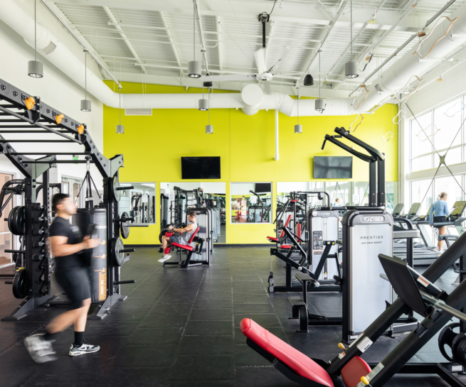 A fitness room with cardio machines, strength equipment, and mirrors reflecting the bright yellow accent wall.