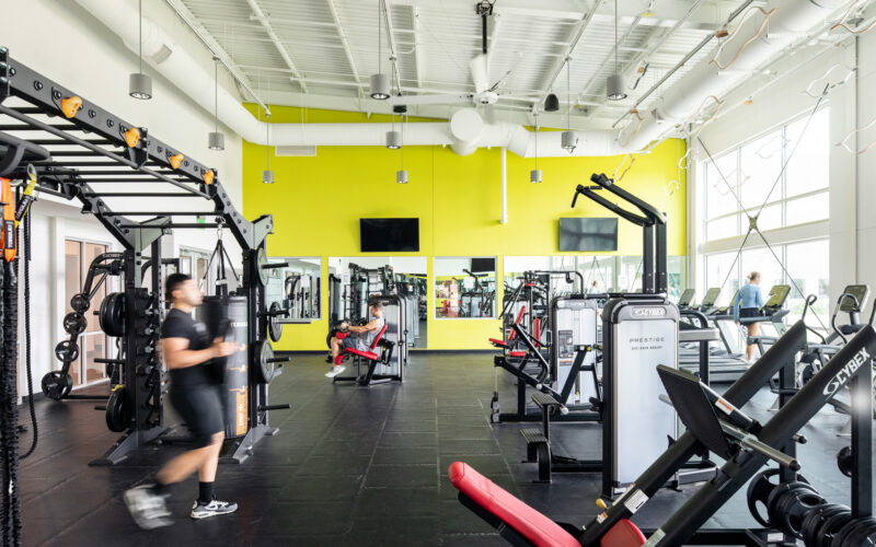 A fitness room with cardio machines, strength equipment, and mirrors reflecting the bright yellow accent wall.