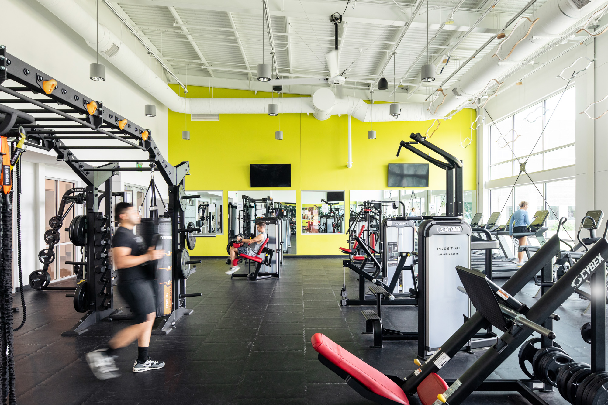 A fitness room with cardio machines, strength equipment, and mirrors reflecting the bright yellow accent wall.