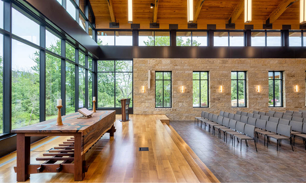 Stage and altar area of Hasley Chapel, with a wooden cross, stone walls, and floor-to-ceiling glass windows overlooking the natural landscape.