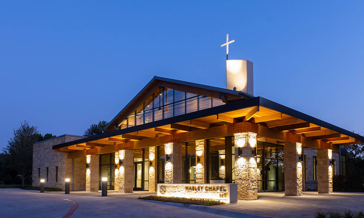 Hasley Chapel's illuminated facade at night, featuring stone columns, wood beams, and large glass windows.