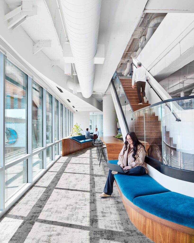 Interior view of GFF’s Victory Park headquarters showing a light-filled corridor with built-in blue velvet seating, floor-to-ceiling curtain wall glazing, and a central stair connecting collaborative workplace levels in a modern open office designed for an architecture firm.