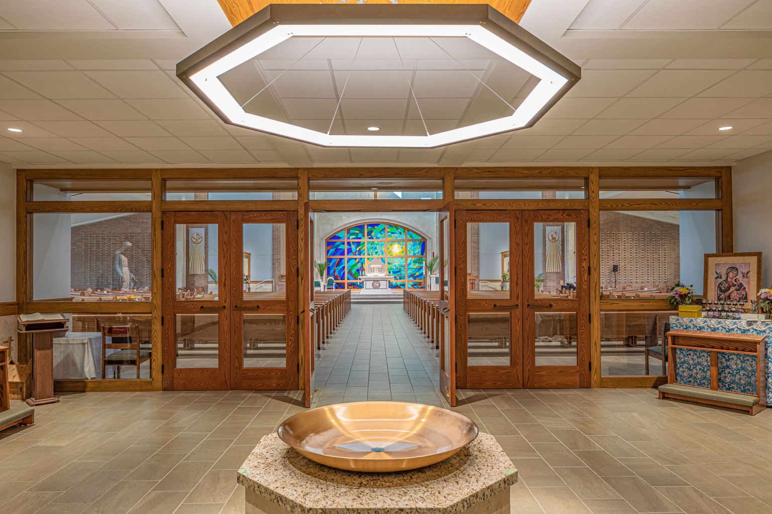 Interior view of the narthex with stained wood-framed doors, a central baptistry, and a glimpse of the nave beyond.
