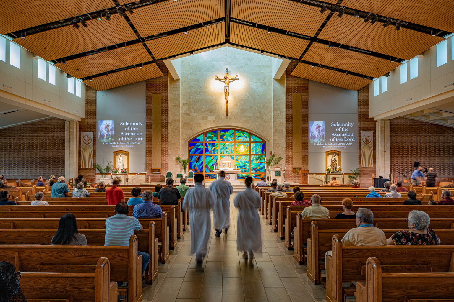 Rear view of the nave showcasing fan-shaped seating, stained glass at the altar, and a warm wood ceiling.