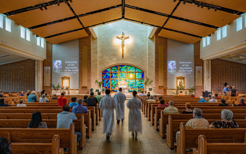 Rear view of the nave showcasing fan-shaped seating, stained glass at the altar, and a warm wood ceiling.