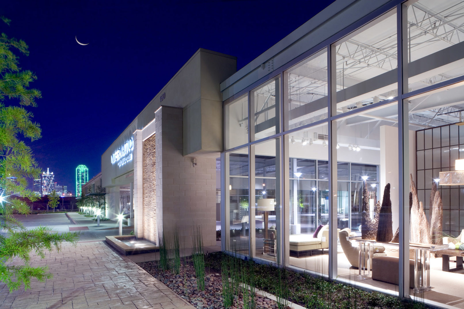 Night view of a glass-fronted showroom with interior lighting and the Dallas skyline visible in the background.